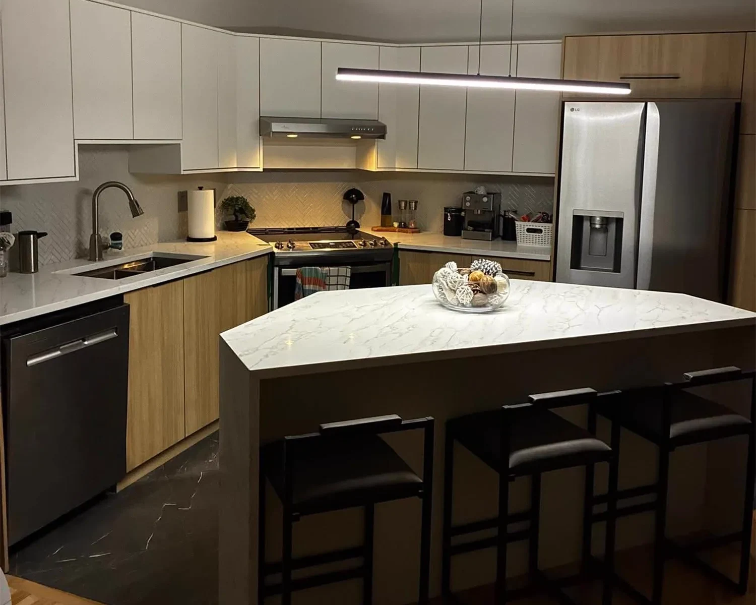 Cuisine éclairée en soirée, îlot en quartz veiné et tabourets noirs, réalisée par Granite Acosta